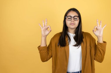 İzole edilmiş sarı arka planda ceket giyen genç ve güzel bir kadın rahatla ve gözün kapalı gülümse parmaklarınla meditasyon hareketi yap.