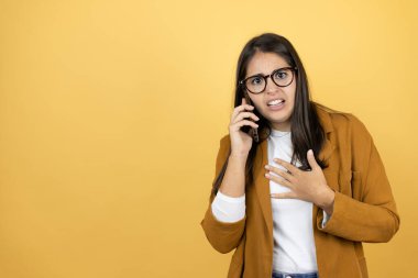 İzole edilmiş sarı arka plan üzerine ceket giyen genç ve güzel bir kadın telefonda endişeli bir ifadeyle konuşuyor.