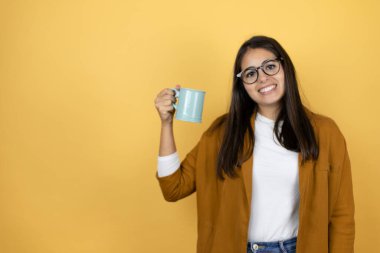 İzole edilmiş sarı arka plan üzerine ceket giyen genç ve güzel bir kadın bir fincan kahve içip eğleniyor.