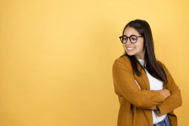 İzole edilmiş sarı arka planda ceket giyen genç ve güzel bir kadın kollarını kavuşturup yan tarafa bakıyor.