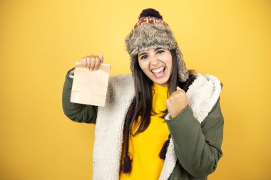 Sarı arka planda yeşil ceketli, şapkalı genç bir kadın elinde bir kese kağıdıyla çok mutlu ve heyecanlı bir şekilde kollarını kaldırıp, gülümseyerek ve başarı için çığlık atarak kazanan bir jest yapıyor.