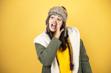 Sarı arka planda şapkası ve yeşil kışlık ceketi olan genç ve güzel bir kadın. Elleri ağızlarında, kötü niyetli konuşmalar fısıldıyor.