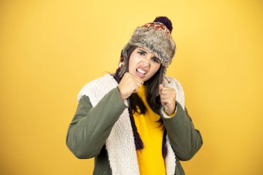 Sarı arka planda şapkası ve yeşil kışlık ceketi olan genç güzel bir kadın dövüşmek için yumruk, agresif ve öfkeli saldırı, tehdit ve şiddet.