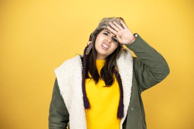 Sarı arka planda şapka ve yeşil kışlık ceket giyen güzel bir kadın. Hastalık ve ateş için alnına dokunuyor. Grip ve soğuk algınlığı için.