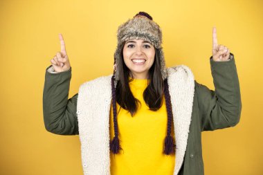 Sarı arka planda yeşil ceketli, şapkalı güzel bir kadın gülümsüyor, kameraya bakıyor ve parmaklarıyla işaret ediyor ve kollarını kaldırıyor.