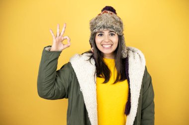 Sarı arka planda yeşil ceketli ve şapkalı güzel bir kadın tamam işareti yapıyor parmaklarıyla ve gülümsemesiyle, mükemmel bir sembol.