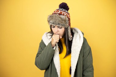 Sarı arka planda yeşil ceketli ve şapkalı güzel bir kadın elleri ağzının yanında öksürüyor.