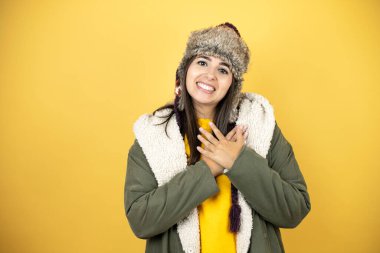 Sarı arka planda yeşil ceketli ve şapkalı güzel bir kadın elleri göğsünde ve yüzünde minnet dolu bir jest ile gülümsüyor..