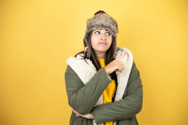 Sarı arkaplan üzerine yeşil bir palto ve şapka takan genç ve güzel bir kadın el ve parmak yanda kafası karışmış bir şekilde işaret ediyor.