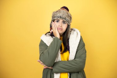 Sarı arka plan üzerinde yeşil bir palto ve şapka giyen genç ve güzel bir kadın çapraz kollardan sıkılmış ve yorgun görünüyor.