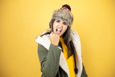 Sarı arka planda yeşil bir palto ve şapka giyen genç ve güzel bir kadın öfkeyle bağırırken kızgın ve kızgın bir şekilde yumruğunu kaldırıyor. Öfke ve agresif kavram.