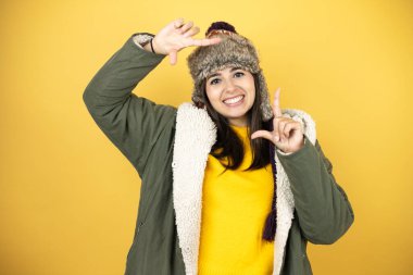 Sarı arka planda yeşil ceketli, şapkalı, güzel bir kadın gülümsüyor ve mutlu yüzlü elleri ve parmakları olan bir çerçeve yapıyor.