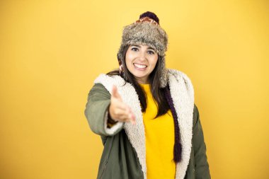 Sarı arka planda yeşil bir palto ve şapka giyen genç ve güzel bir kadın selamlama ve hoş geldin demek için gülümseyerek el sıkışır.