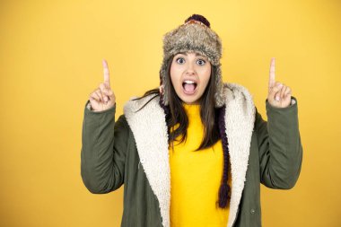Sarı arka planda yeşil bir palto ve şapka giyen genç ve güzel bir kadın kameraya bakıp parmaklarını kaldırıp kollarını kaldırdığında şaşırmış ve şaşırmıştı.