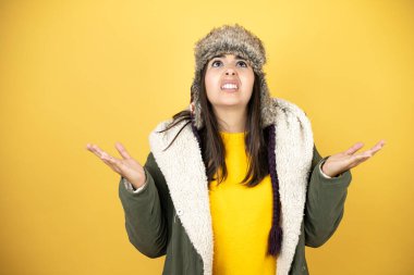 Sarı arka planda yeşil bir palto ve şapka giyen genç ve güzel bir kadın. Kolları ve elleri havada, kafası karışmış bir ifade.