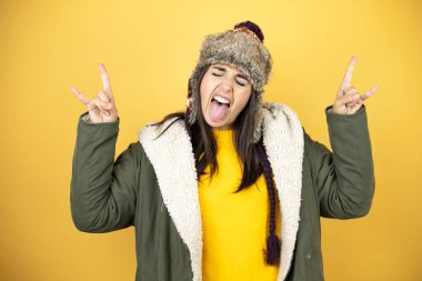Sarı arka planda yeşil bir palto ve şapka giyen genç ve güzel bir kadın çılgın bir ifadeyle elleri havada rock sembolü yapıyor.