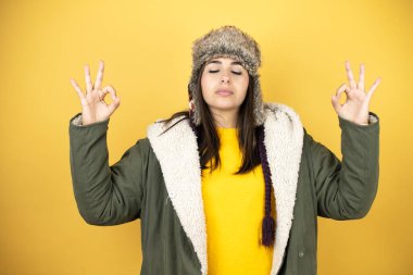 Sarı arka planda yeşil ceketli ve şapkalı güzel bir kadın rahatla ve gözleri kapalı gülümse. Parmaklarıyla meditasyon hareketi yap.