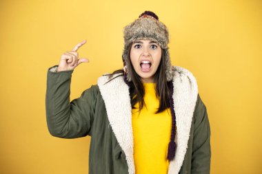 Sarı arka planda yeşil bir palto ve şapka giyen genç ve güzel bir kadın gülümsüyor ve el hareketleriyle parmaklarıyla küçük boy işareti yapıyor. Ölçü kavramı.