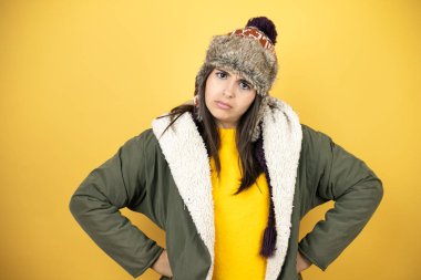 Sarı arka plan üzerine yeşil bir palto ve şapka giyen genç ve güzel bir kadın şüpheci ve gergin, yüzünde onaylamayan bir ifade var.