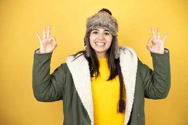 年轻美丽的女人 头戴一顶帽子 身穿绿色冬衣 背景是黄色的 手握手指 象征着完美 — 图库照片