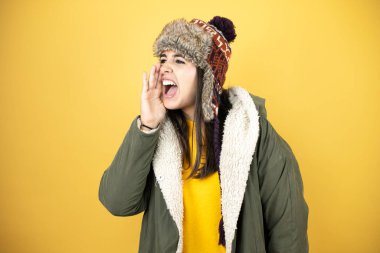 Sarı arka planda yeşil bir palto ve şapka giyen genç ve güzel bir kadın elleri ağızda bağırarak ve yüksek sesle bağırıyor.