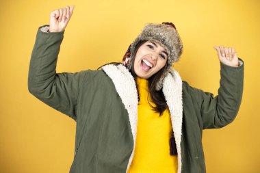 Sarı arka planda yeşil ceketli ve şapkalı güzel bir kadın. Çok mutlu ve heyecanlı. Kollarını kaldırmış, gülümseyen ve başarı için çığlık atan bir jest yapıyor..