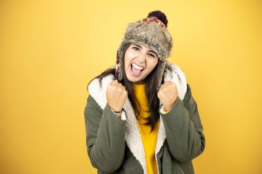 Sarı arka planda yeşil ceketli ve şapkalı güzel bir kadın. Çok mutlu ve heyecanlı. Kollarını kaldırmış, gülümseyen ve başarı için çığlık atan bir jest yapıyor..