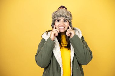 Sarı arka planda şapkalı ve yeşil ceketli güzel bir kadın gülümsüyor ve parmaklarıyla ve ağzıyla işaret ediyor.