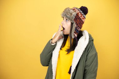 Genç, güzel bir kadın, sarı arka planda yeşil bir palto ve şapka takıyor. Eli ağzında ve şaşırmış, yan bakıyor.