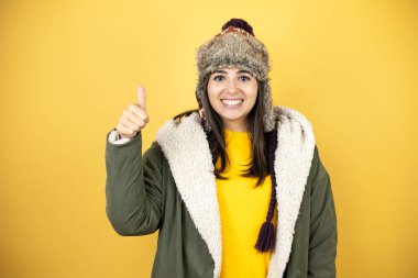 Sarı arka planda yeşil ceketli, şapkalı güzel bir kadın gülümsüyor ve baş parmaklarıyla işaret veriyor.