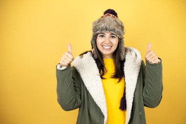 Sarı arka planda yeşil ceketli, şapkalı güzel bir kadın gülümsüyor ve baş parmaklarıyla işaret veriyor.