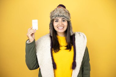 Sarı arka planda yeşil ceketli, şapkalı güzel bir kadın gülümsüyor ve elinde beyaz bir kart tutuyor.