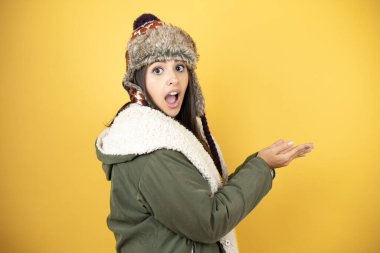 Sarı arka planda yeşil bir palto ve şapka giyen güzel bir kadın ellerinde boş bir alan olan bir şeyi tutarak bir şey yapıştırıyor.