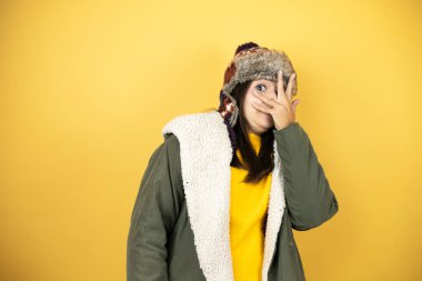 Sarı arka plan üzerinde şapka ve yeşil kışlık ceket giyen genç güzel bir kadın. Şok içinde yüzünü ve gözlerini kapayarak utangaç bir ifadeyle parmaklarının arasından bakıyor.