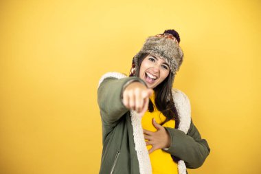 Sarı arka planda yeşil ceketli, şapkalı genç ve güzel bir kadın sana gülüyor, kamerayı işaret ediyor, vücudunu gösteriyor, utanç ifadesi var.