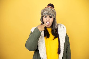 Sarı arka planda, şapkası ve yeşil kışlık ceketi olan genç ve güzel bir kadın. Eli ağzının içindeyken iğreniyor.