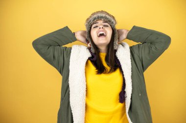 Sarı arka planda yeşil ceketli, şapkalı, güzel bir kadın rahatlıyor ve esniyor, kolları ve elleri başının arkasında ve boynunun arkasında gülümsüyor.