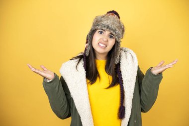 Sarı arka planda yeşil bir palto ve şapka giyen genç ve güzel bir kadın. Kolları ve elleri havada, kafası karışmış bir ifade.