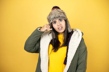 Sarı arka planda yeşil ceketli, şapkalı genç ve güzel bir kadın telefonda kafası karışmış bir şekilde el ve parmaklarıyla telefon jesti yapıyor.