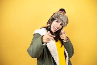 Sarı arka planda yeşil ceketli, şapkalı genç ve güzel bir kadın seni ve kamerayı işaret ediyor, mutlu ve mutlu gülümsüyor.