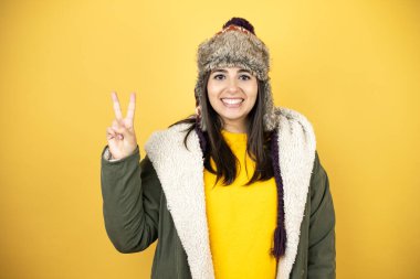 Sarı arka planda yeşil ceketli ve şapkalı güzel bir kadın iki numaralı parmaklarını gösterirken kendinden emin ve mutlu bir şekilde gülümsüyor.