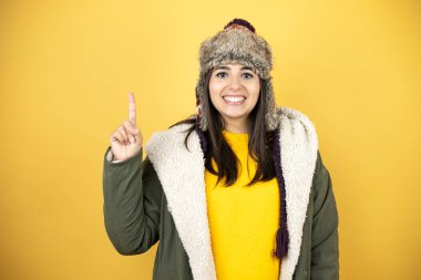 Sarı arka planda yeşil bir palto ve şapka giyen genç ve güzel bir kadın bir numaralı parmaklarını gösterirken kendinden emin ve mutlu bir şekilde gülümsüyor.