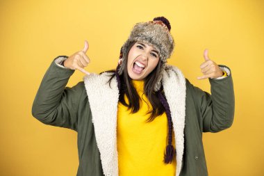 Sarı arka planda yeşil bir palto ve şapka giyen genç ve güzel bir kadın çılgın bir ifadeyle elleri havada rock sembolü yapıyor.