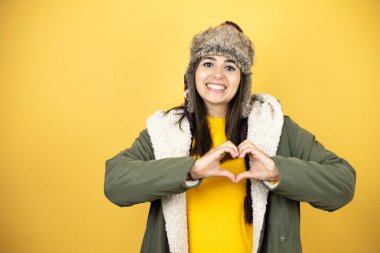 Sarı arka planda yeşil bir palto ve şapka giyen genç ve güzel bir kadın. Aşkla gülümsüyor. Elleriyle kalp sembolü ve şekli gösteriyor.