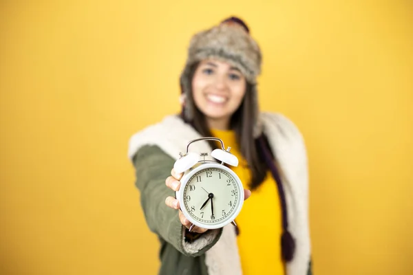 帽子をかぶっている若い美しい女性と前に時計を示す黄色の背景に緑色の冬のコート — ストック写真