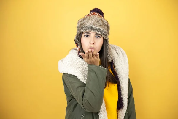 年轻美丽的女人戴着一顶帽子 披着绿色的冬衣 背景是黄色的 看着摄像机 用手与空气亲吻 是可爱而性感的 爱的表达 — 图库照片