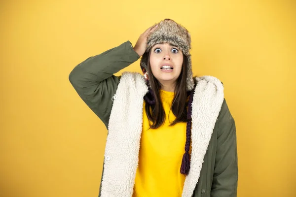 年轻而漂亮的女人 戴着一顶帽子 披着绿色的冬季外套 披着黄色的背景 一只手捂着头笑着 好像忘了什么似的 — 图库照片