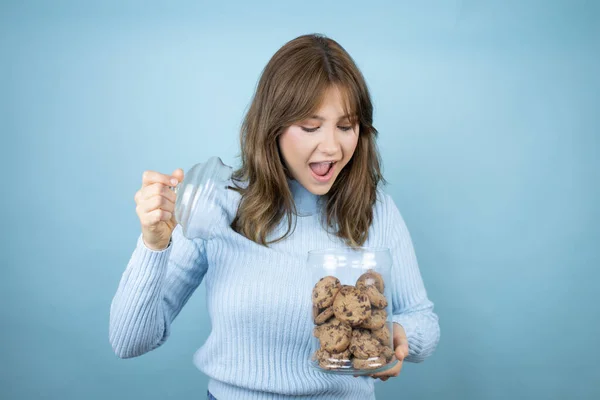Young beautiful woman holding chocolate chips cookies jar over isolated blue background amazed, opening and looking at the cookie jar