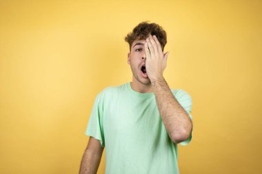 Sarı arka planda yeşil bir tişört giyen yakışıklı bir adam bir gözünü eliyle kapatıyor, yüzünde kendine güvenen bir gülümseme ve sürpriz bir duygu var..