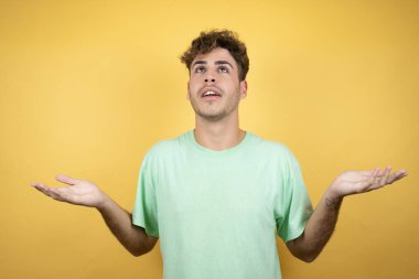 Sarı arka planda yeşil bir tişört giyen yakışıklı bir adam. Hiçbir fikri olmayan, elleri ve kolları havada, kafası karışmış bir adam.